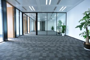 A large and clean office space with light grey carpet tiles. The carpet tiles are square shaped and laid in a grid pattern. A potted plant sits in the corner of the office next to a glass mounted on the wall.