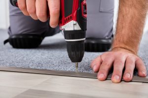 A person kneels while using a power drill to secure a transition strip between carpet tiles and hardwood flooring, demonstrating practical carpet tile design ideas and installation techniques.